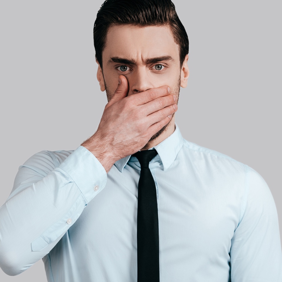Trying to stop gossiping. Surprised young man in white shirt and tie covering mouth with hand and looking at camera while standing against grey background