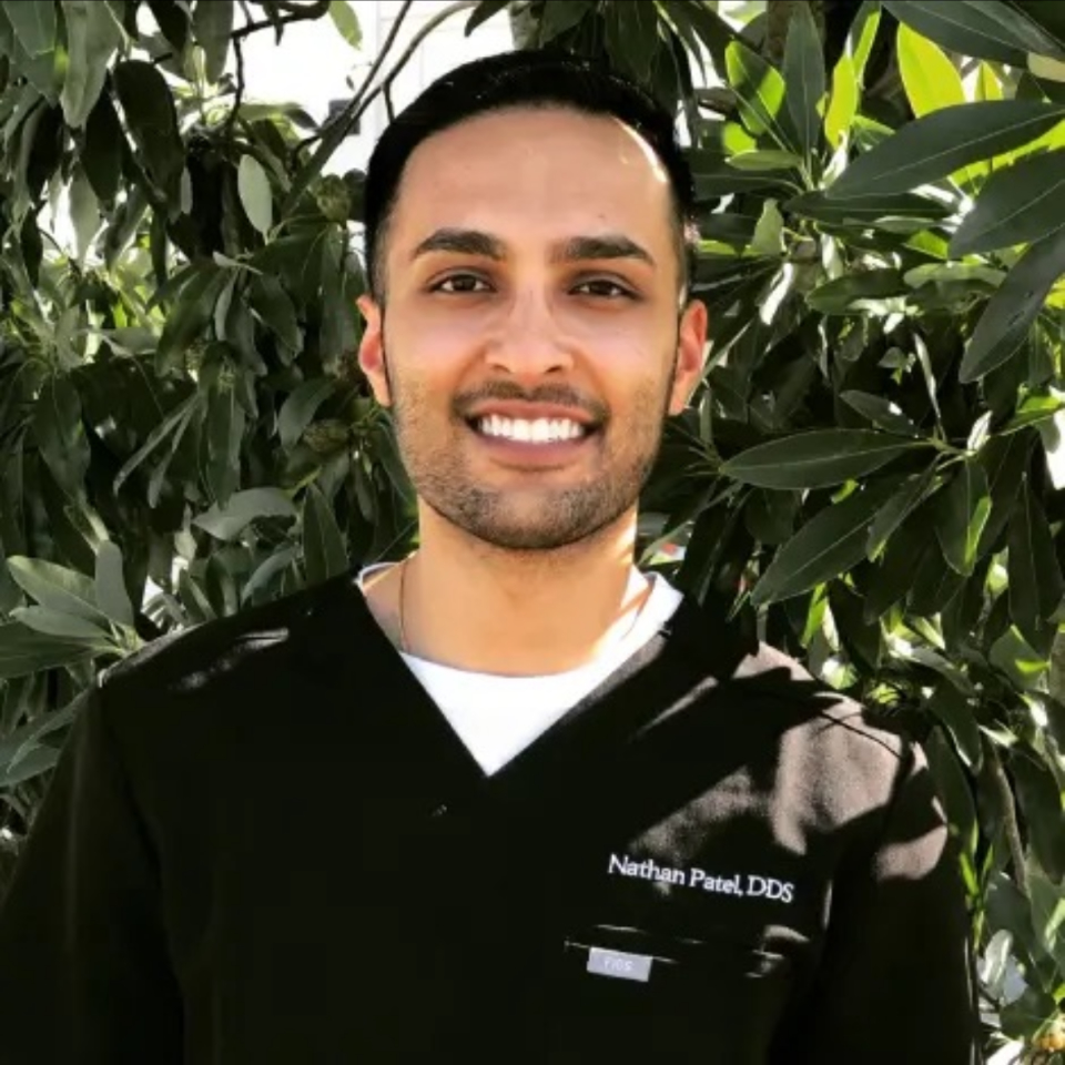 A person wearing black medical scrubs with the name "Nathan Patel, DDS" stands outdoors in front of leafy green foliage, smiling at the camera.
