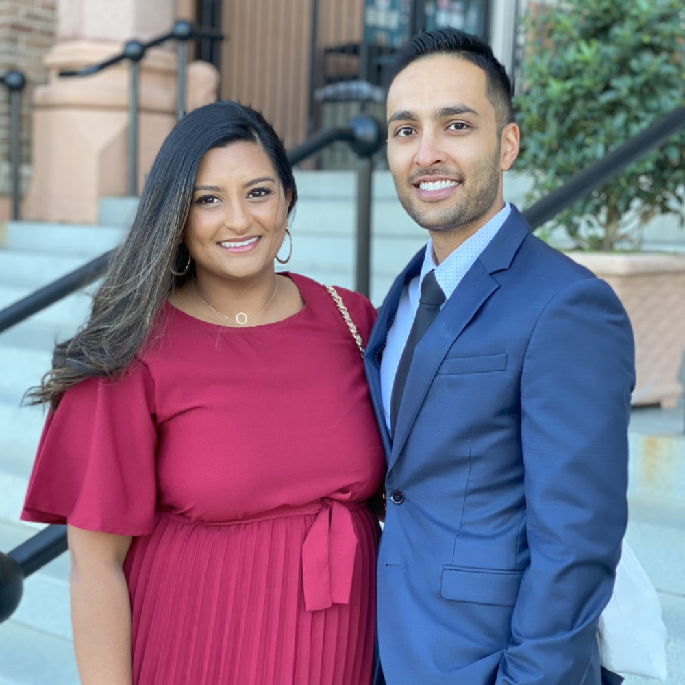 Two people stand side by side outdoors, smiling at the camera. The woman wears a maroon dress and the man wears a blue suit. Steps and a potted plant are visible in the background.