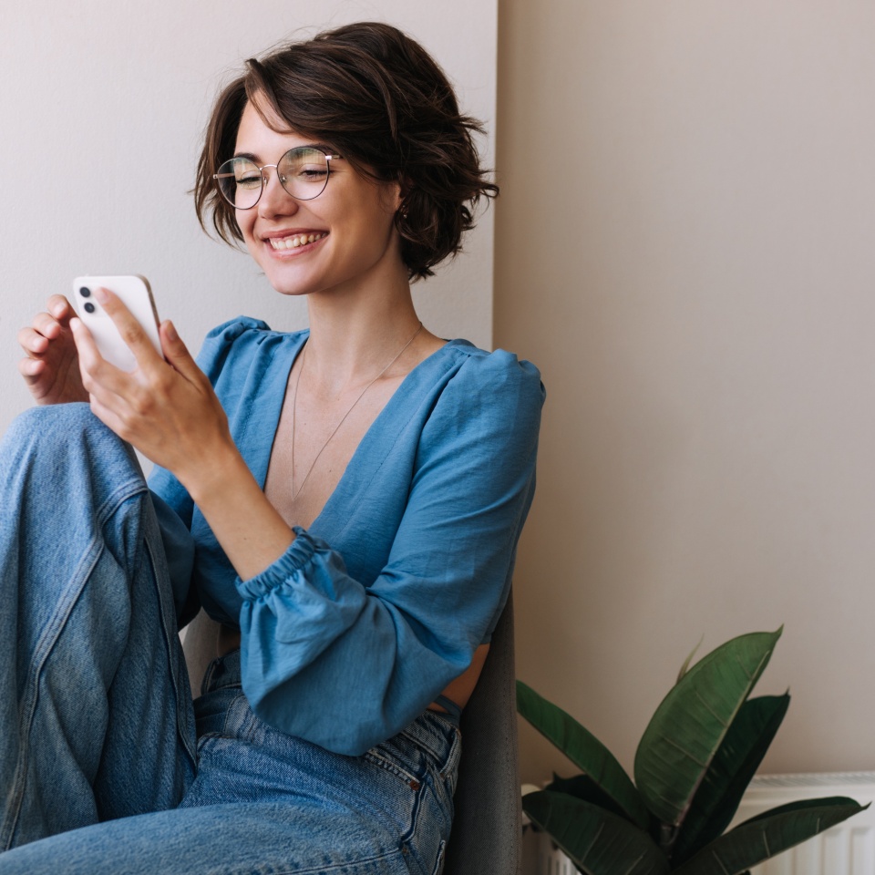Charming woman holding phone at home . Caucasian smiling brunette woman looking and chatting on smartphone sitting on the floor. Concept of lifestyle, use technology
