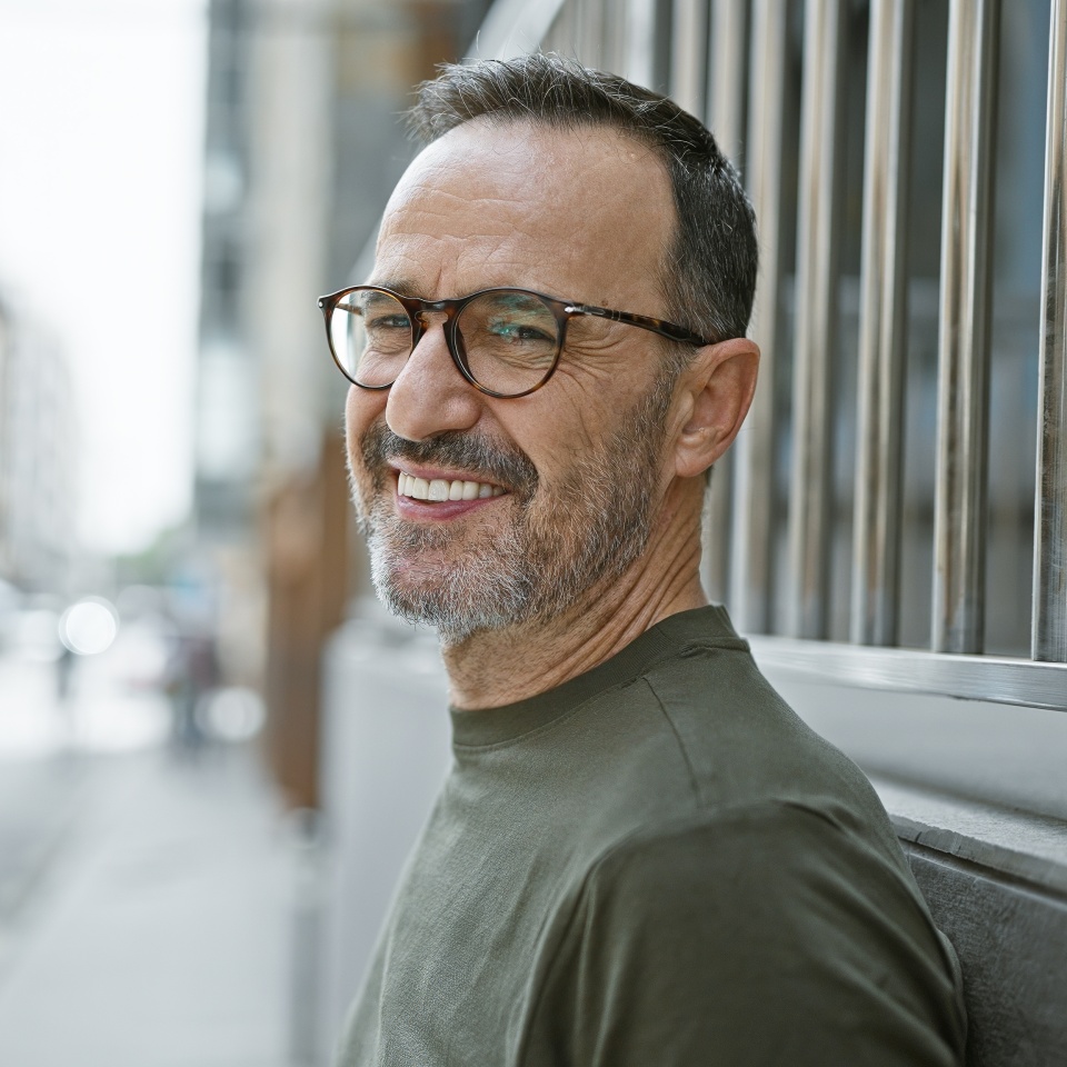Brimming with joy, middle age man with grey hair stands confidently on city street, his smile beaming happiness as he subtly laughs, his mature expression telling tales of a fulfilled life.