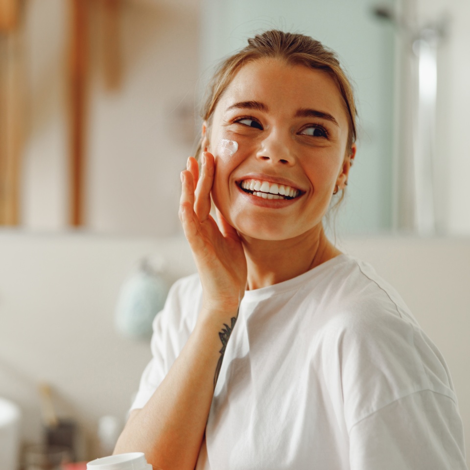 Happy woman applying hydrating moisturizer on her face sitting in bathroom. Home beauty routine